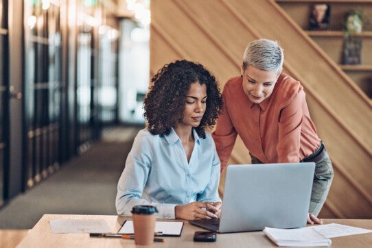 Two business womens working on laptop talking showing discuss some work. Desk full of documents, coffee, telephone and pens modern work space. Horizontal photo.