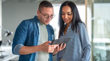 A male and female Hispanic duo engaged in work on a digital tablet, dressed in smart casual office attire, within a modern workspace.