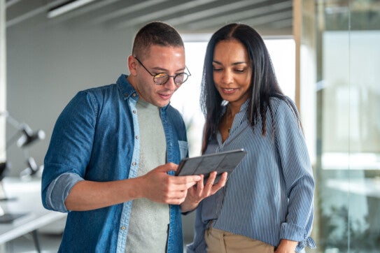 A male and female Hispanic duo engaged in work on a digital tablet, dressed in smart casual office attire, within a modern workspace.